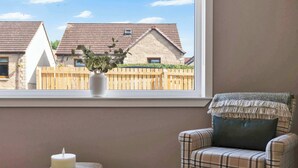 Ein Zimmer mit Blick auf Häuser durch ein Fenster, ein Sessel mit kariertem Überwurf und eine Kerze auf einem Tablett.
