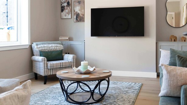 Ein Wohnzimmer mit einem karierten Sofa, einem runden Couchtisch mit einer Kerze und einem an der Wand befestigten Flachbildfernseher.