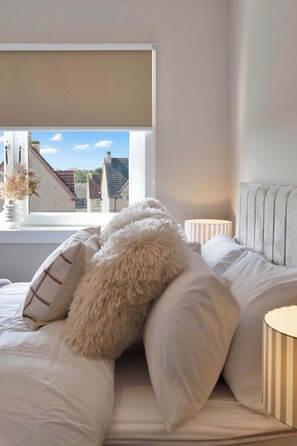 A bedroom with a bed, pillows, a window with a view of buildings, and a bedside lamp.