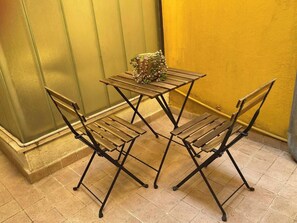 Ein kleiner Außenbereich mit einem Metalltisch und -stühlen, einer Blumenkomposition auf dem Tisch und einer gelben Wand im Hintergrund.