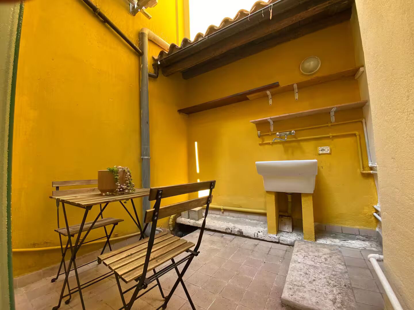 A small outdoor area with a wooden bench, a table with a vase, and a yellow wall.