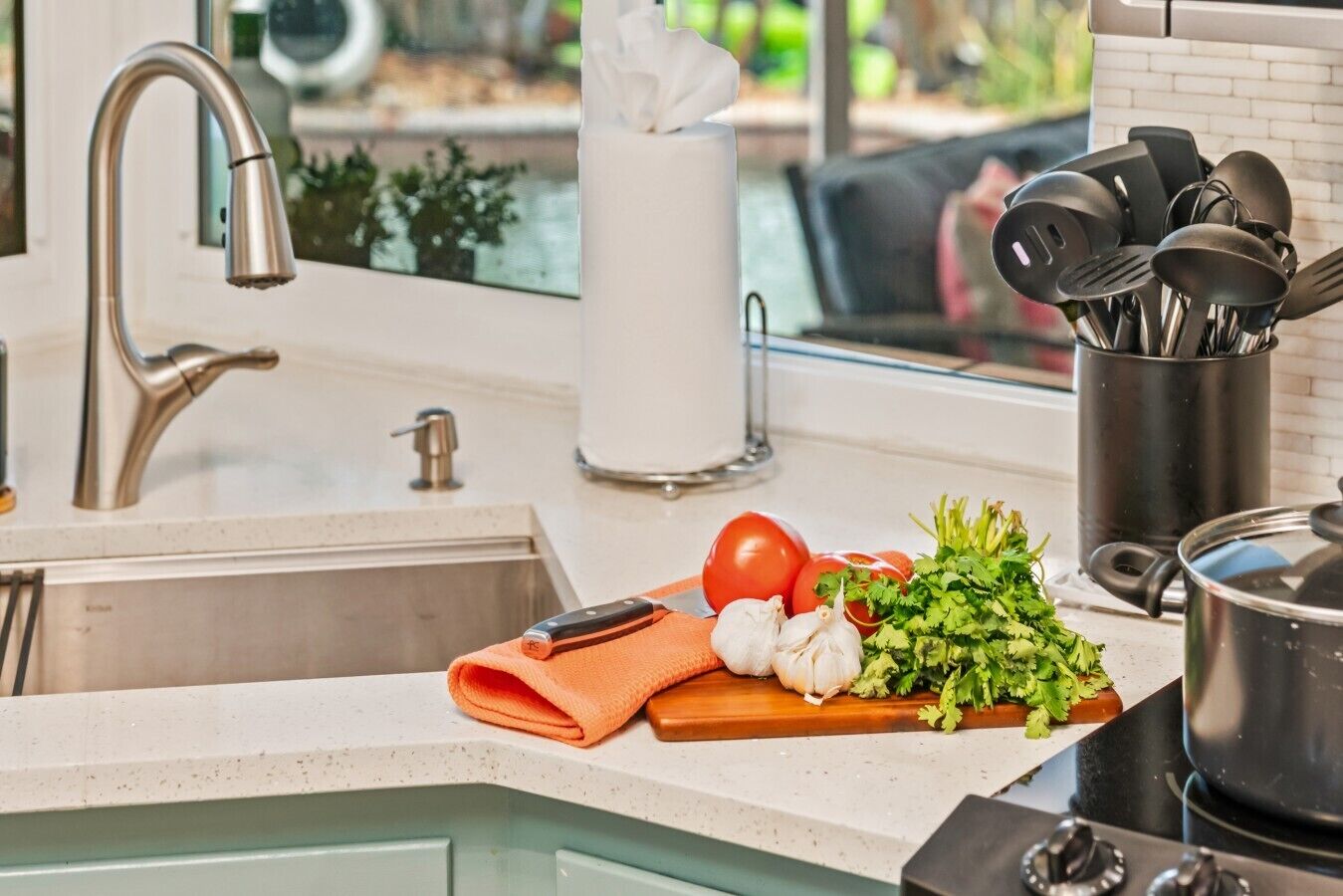 Una cocina con un grifo de acero inoxidable, tabla de cortar con verduras, olla y un recipiente para utensilios.