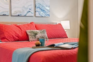 Een bed met rode beddengoed en een kussen van palmblad, een nachtkastje met een lamp, en abstracte wanddecoratie.