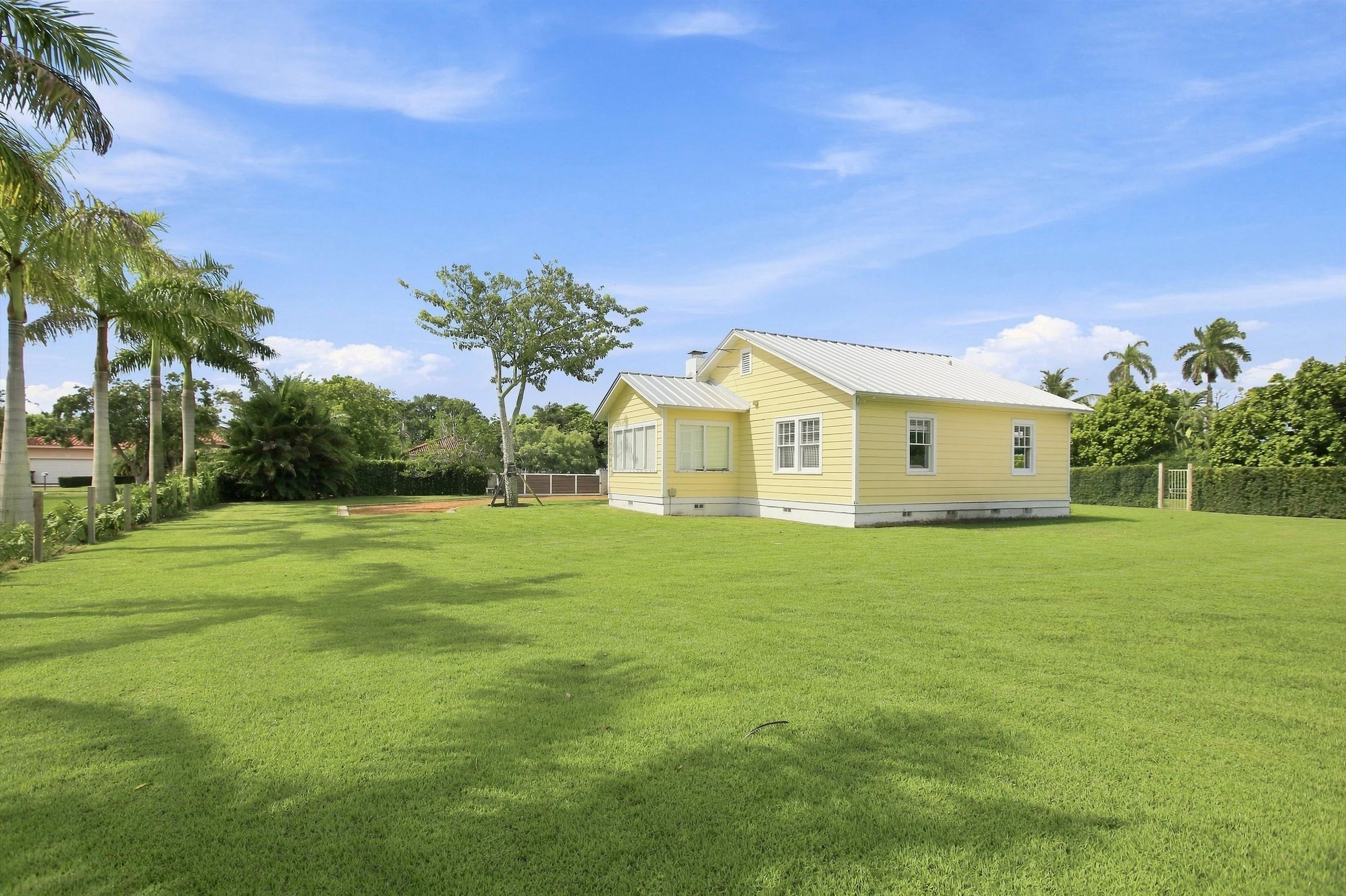 A single-story yellow house with a white roof, surrounded by a well-maintained lawn and palm trees.