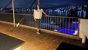 Eine Person steht auf einem Balkon mit Geländer und blickt auf ein Gewässer und eine nächtliche Stadtansicht.