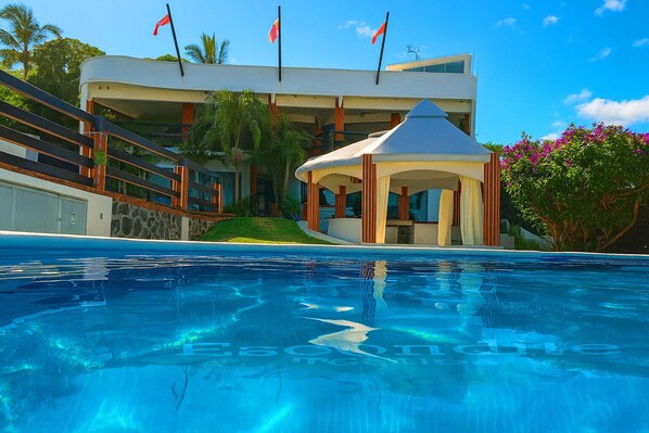 A pool with a gazebo and a building with multiple flags. - Escondite Teques / Alberca climatizada, diversión y espacio para grupos (Tequesquitengo)
