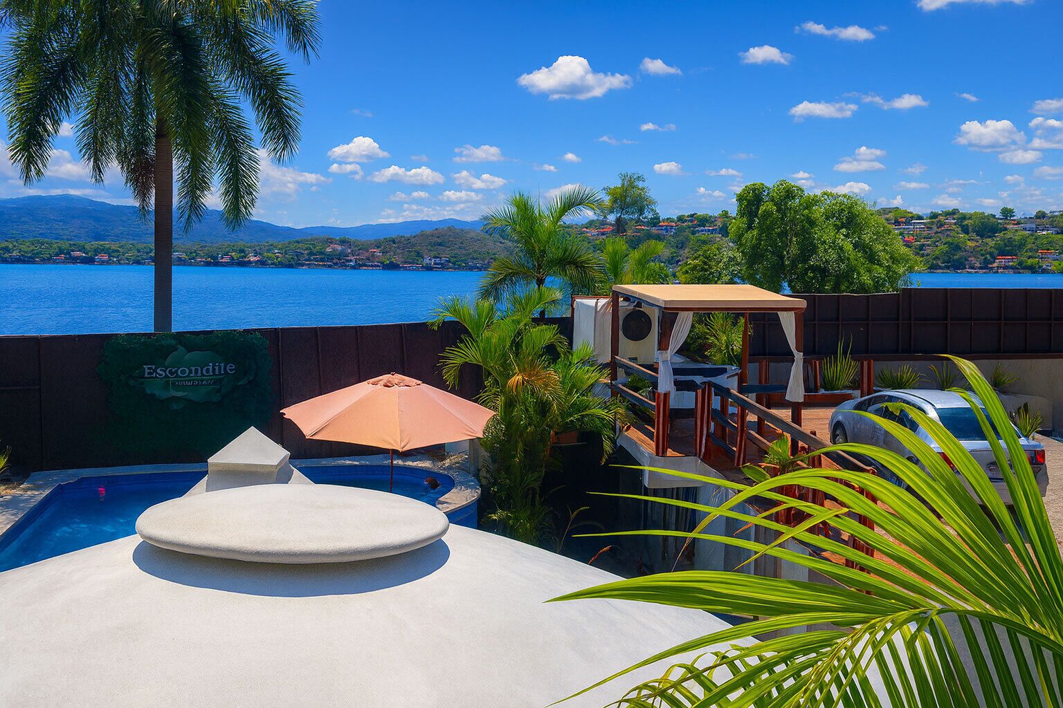 A pool area with a view of a body of water and hills, a covered seating area, and a sign for 'Escondite'.