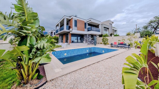 A modern two-story house with a swimming pool, surrounded by tropical plants.