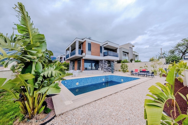 A modern two-story house with a swimming pool, surrounded by tropical plants.