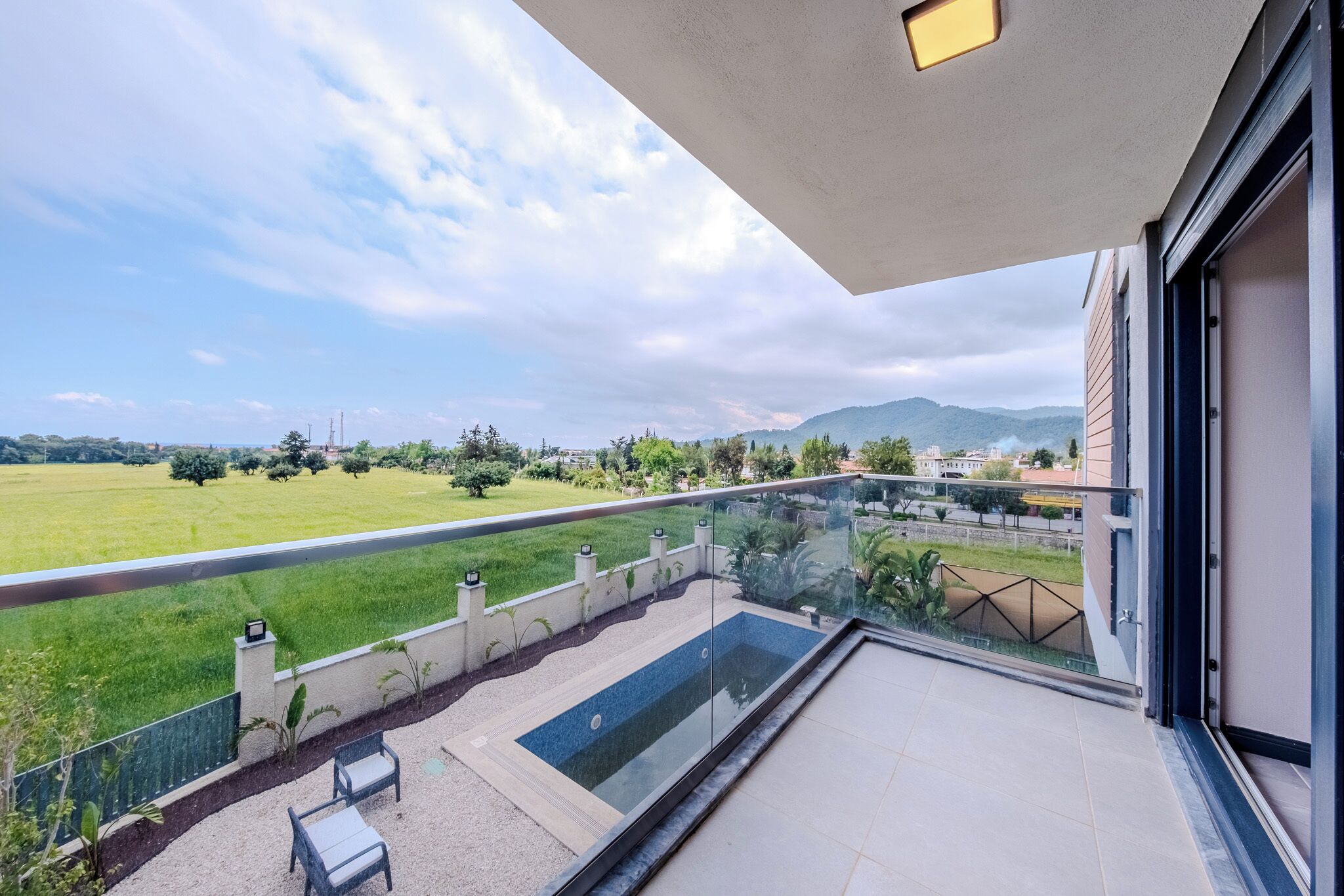A balcony with a glass barrier, a pool, and a view of a green landscape and distant hills.