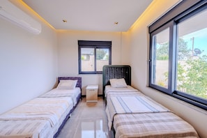 A room with two single beds, a small bedside table, a window, and an air conditioning unit.