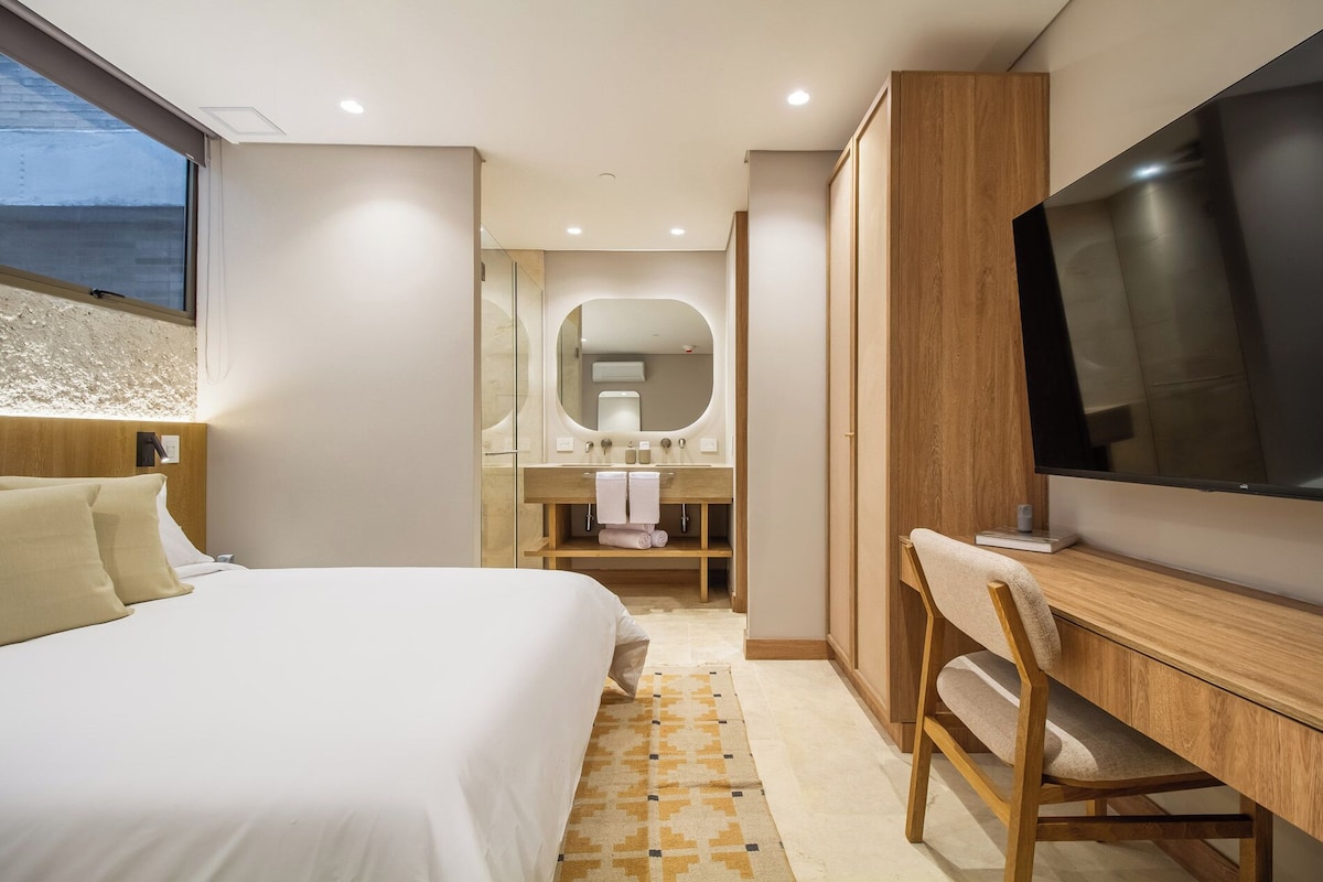 A hotel room with a bed, a desk with a chair, a TV, a bathroom with a mirror, and a window.