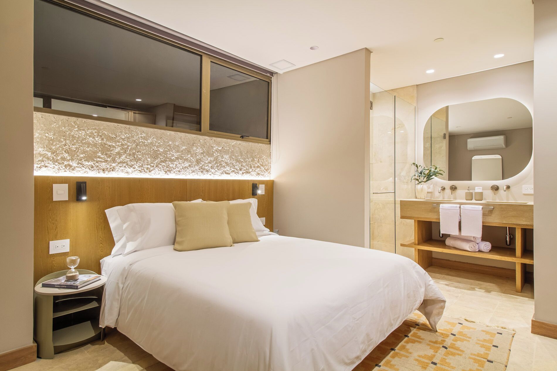 Habitación de hotel con una cama grande, cabecera de madera, mesita de noche, tocador, espejo, baño con lavamanos y perchero.