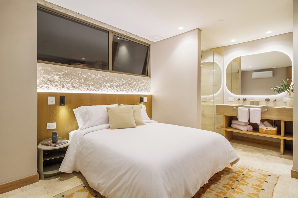 A hotel room with a large bed, a bedside table, a mirror, and a bathroom with a vanity.