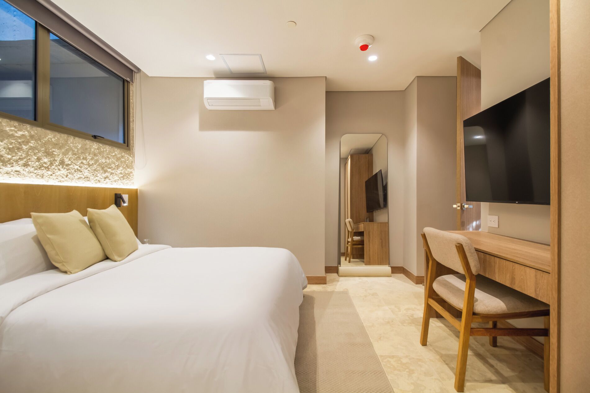 A hotel room with a bed, a desk, a chair, a television, and an air conditioning unit.
