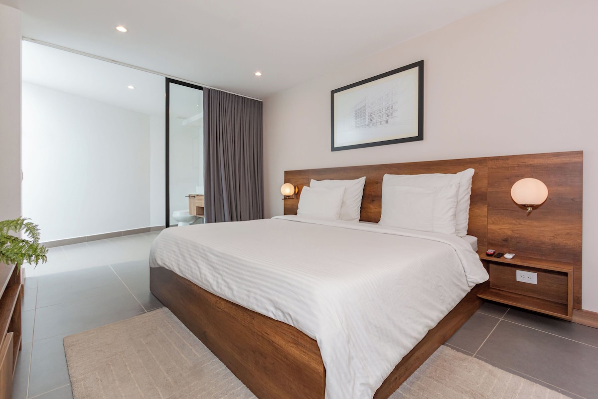 A modern hotel room with a large bed, a wooden headboard, a bedside table with a lamp, a framed picture on the wall, and a view of the outside through a window.