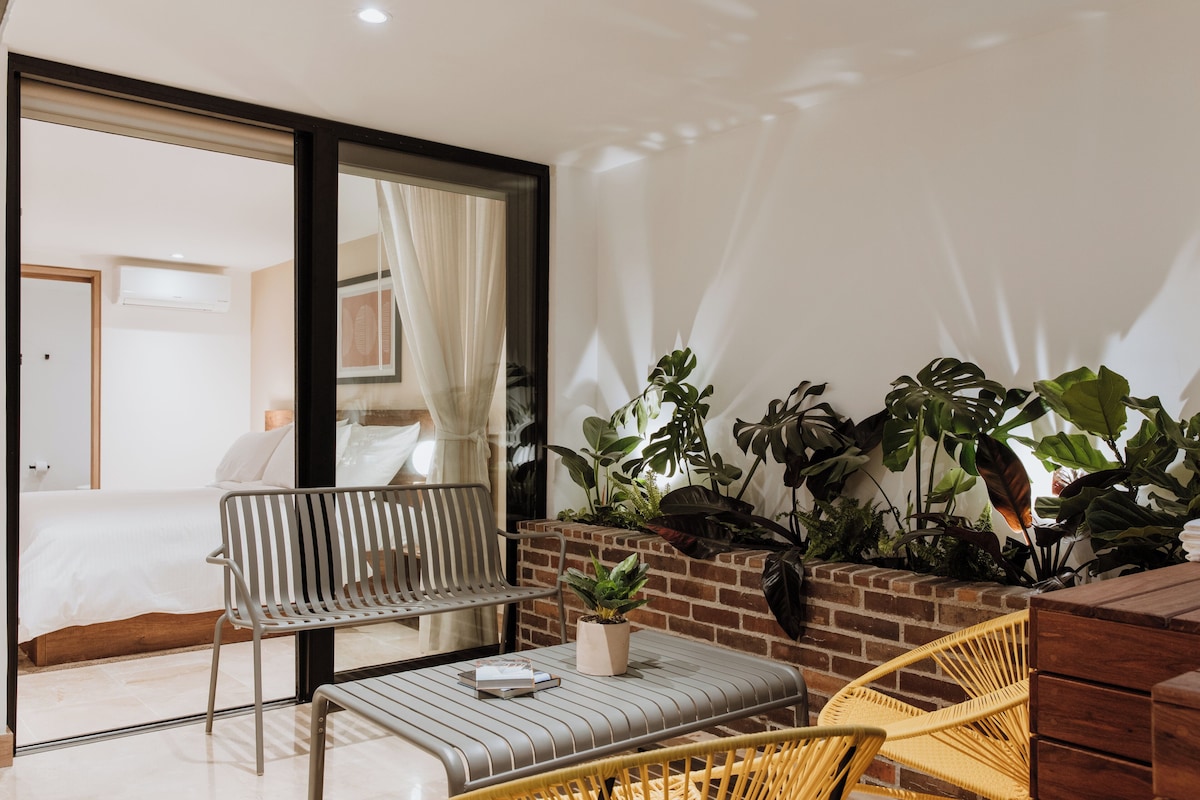 A modern hotel room with a balcony, a bed, a seating area with a table and chairs, and a variety of plants.