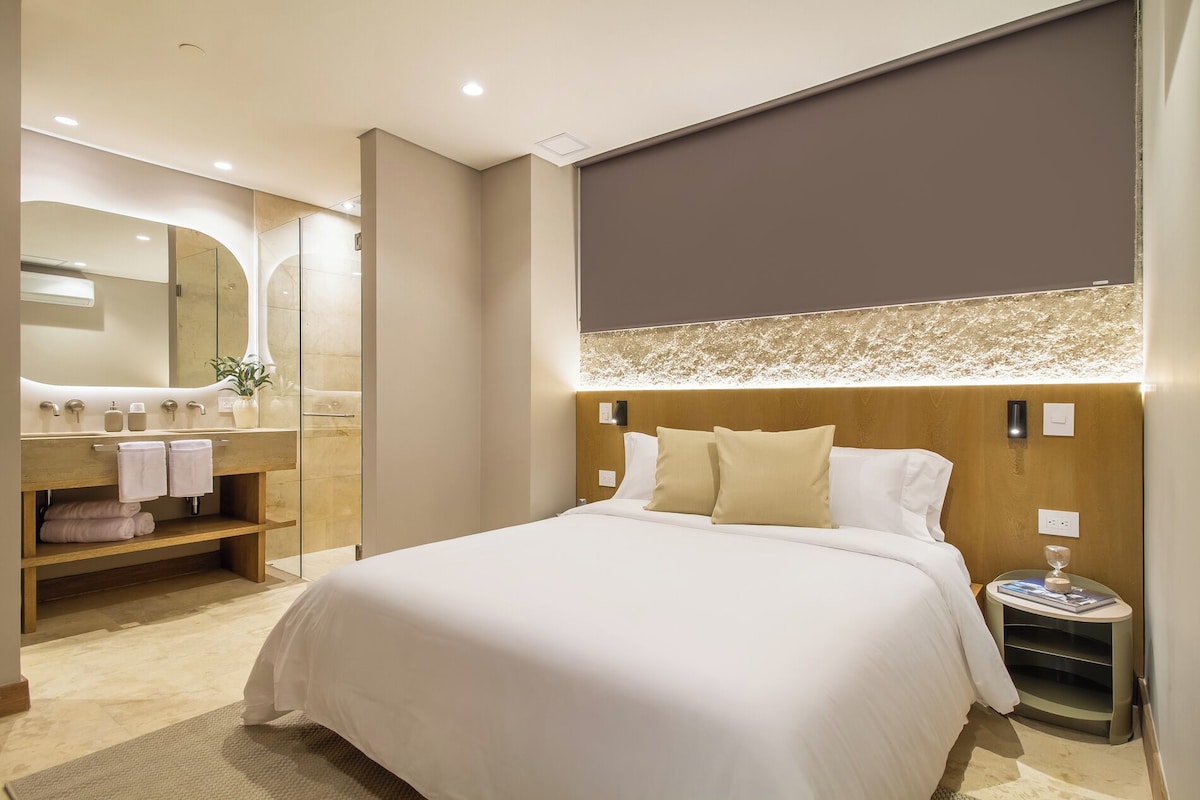 A hotel room with a large bed, a wooden headboard, a bedside table with a glass, and a bathroom with a mirror and sink.