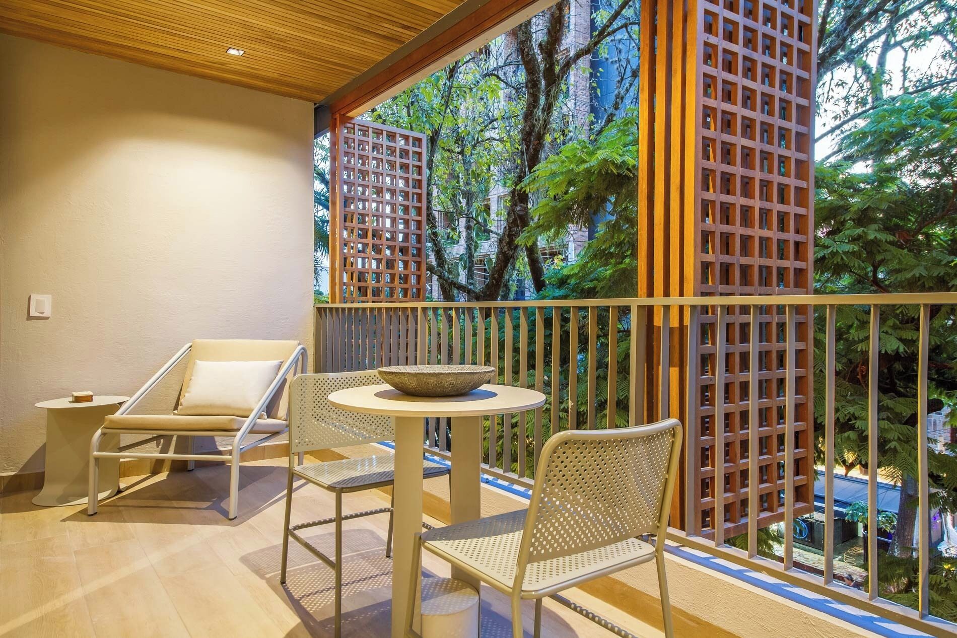 A balcony with two chairs and a table, a wooden ceiling, and a view of trees and buildings.