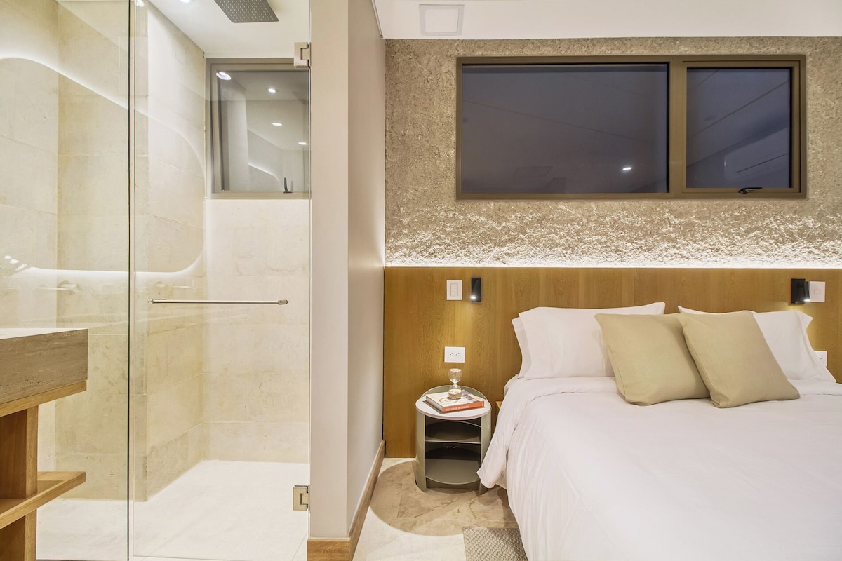 A modern hotel room with a glass shower enclosure, a wooden headboard, and a bedside table with a book and a glass.