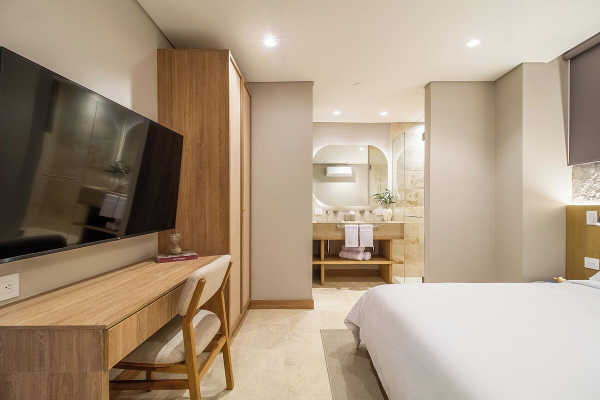 A modern hotel room with a large flat-screen TV, a wooden desk, a comfortable chair, a large bed, a bathroom with a mirror, and a shower.