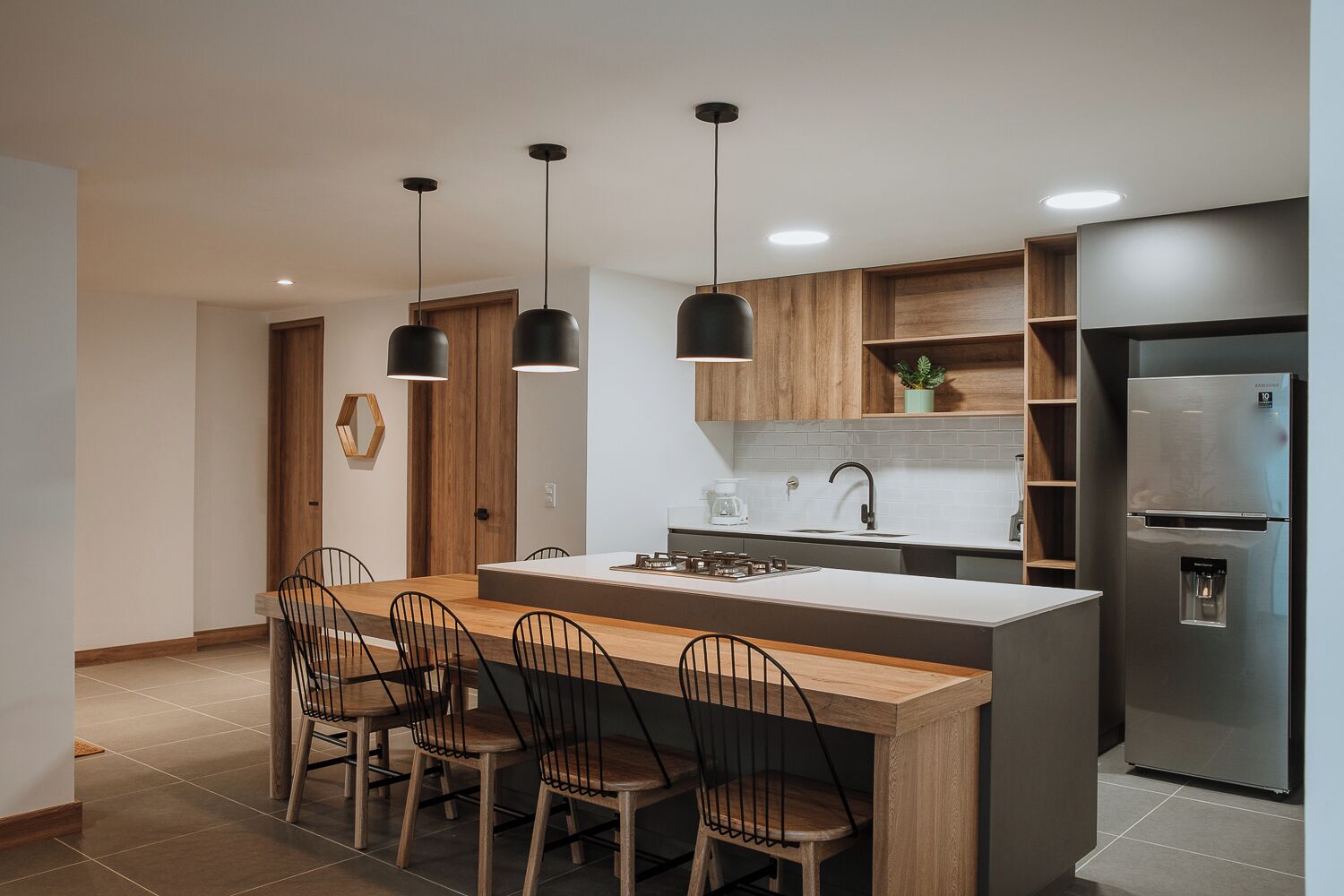 Una cocina moderna con una isla central, taburetes de bar, luces colgantes, un refrigerador integrado y armarios de madera.