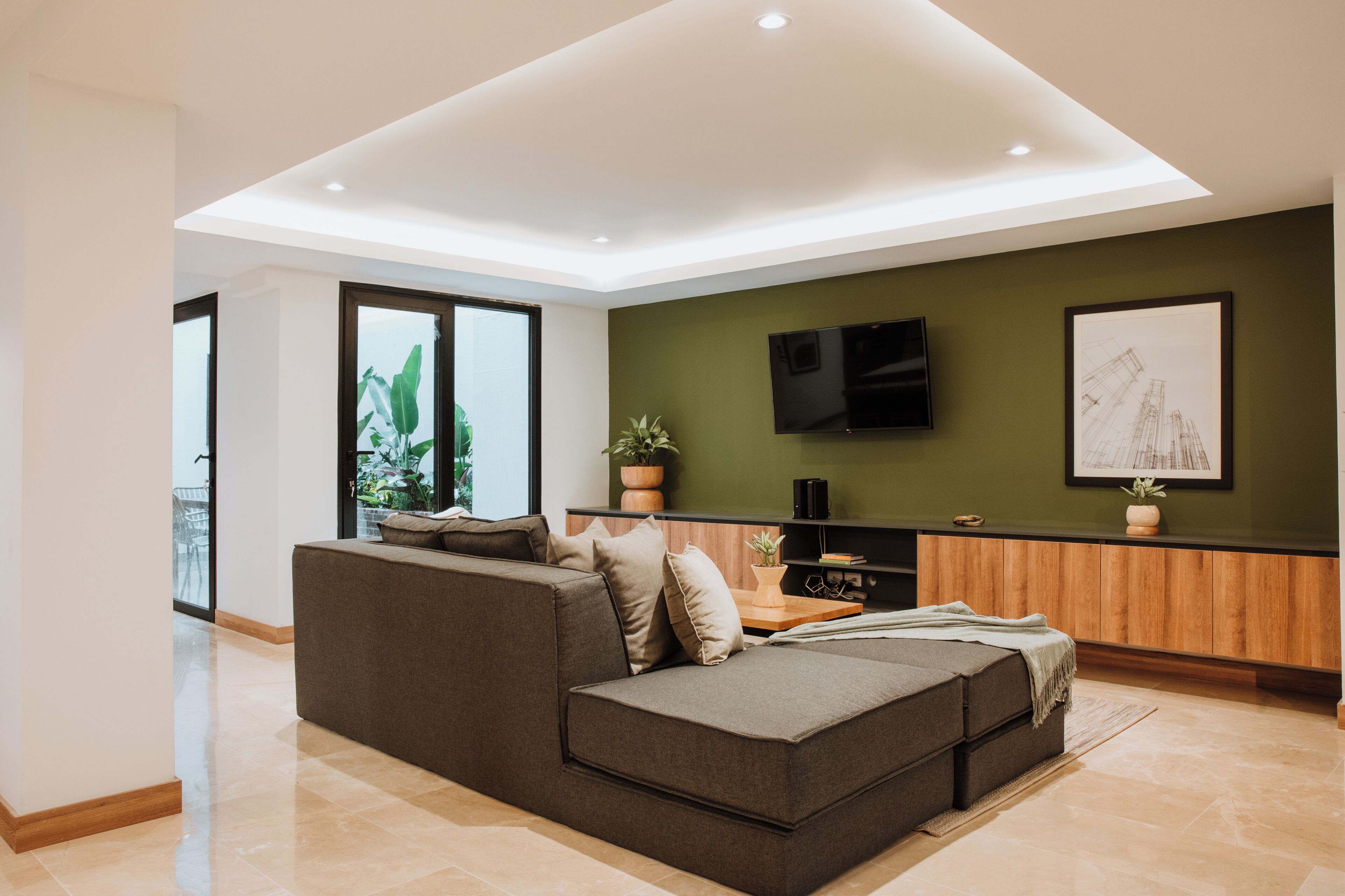 Una sala de estar moderna con un sofá seccional, un televisor de pantalla plana montado en la pared y un ventanal con vistas al exterior.