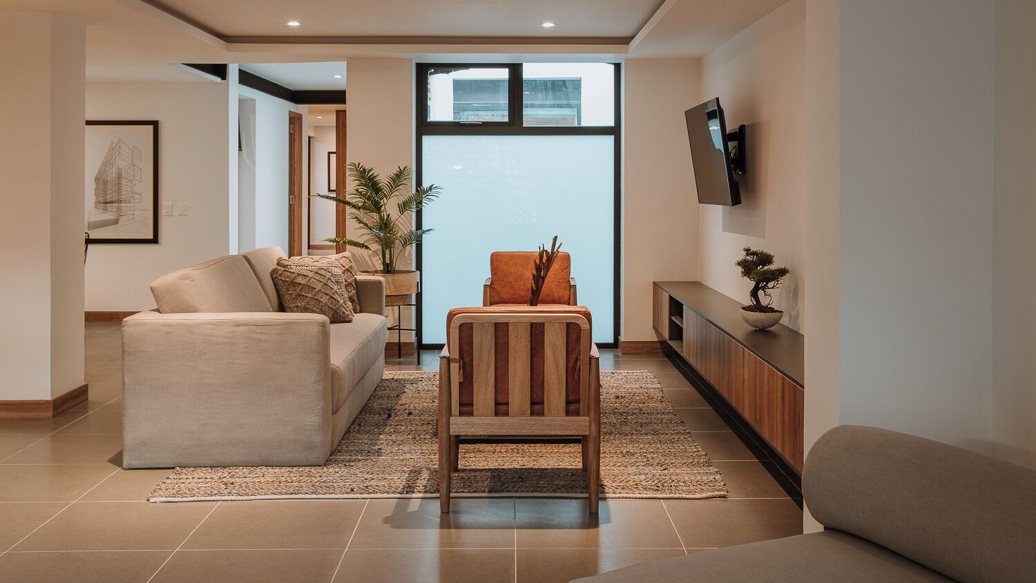 A modern living room with a sofa, a wooden chair, a TV mounted on the wall, and a potted plant.