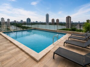 A rooftop swimming pool with a city skyline view, surrounded by a glass railing and furnished with lounge chairs. - Fully equipped apartment with balcony, pool, and gym. (Barueri)