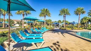 A poolside area with blue lounge chairs and umbrellas, surrounded by palm trees and a clear blue sky.