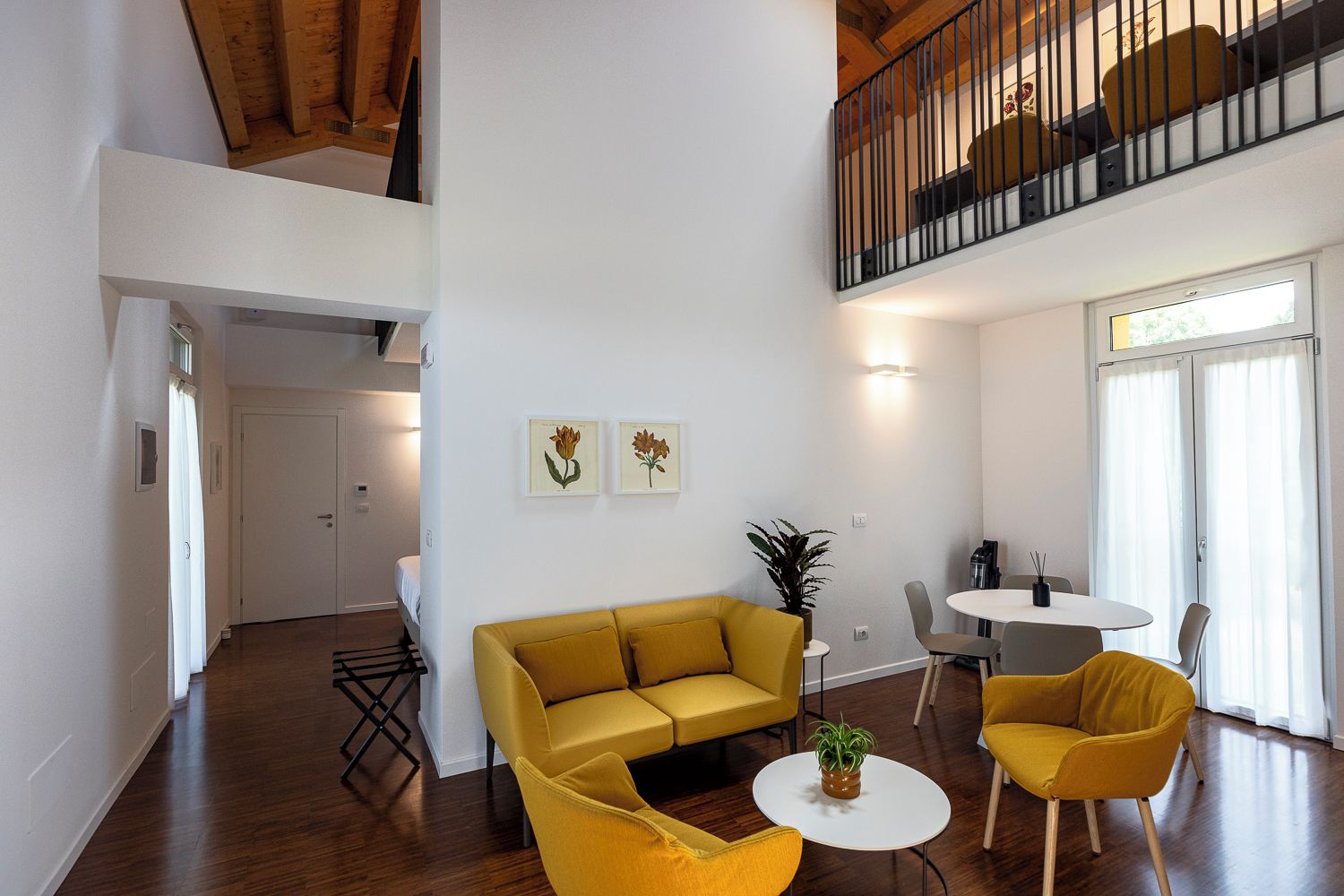 Un salón amplio con un sofá amarillo, una mesa redonda y una mesa de comedor con sillas. Se ve una escalera que sube a un nivel superior.