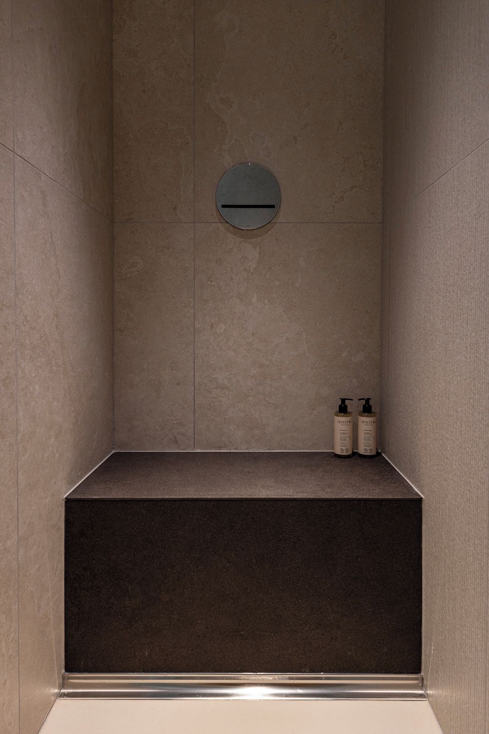 A minimalist bathroom with a dark floor, a round mirror, and two toiletry bottles on the wall.
