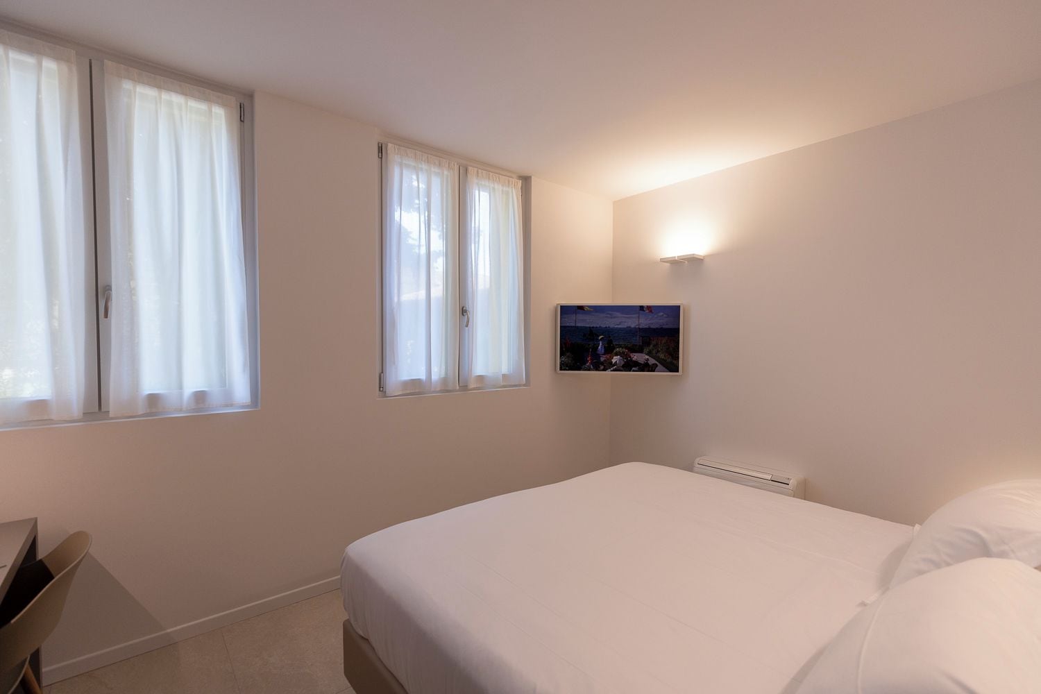 A bedroom with a bed, a chair, a nightstand, a wall-mounted TV, and a window with curtains.