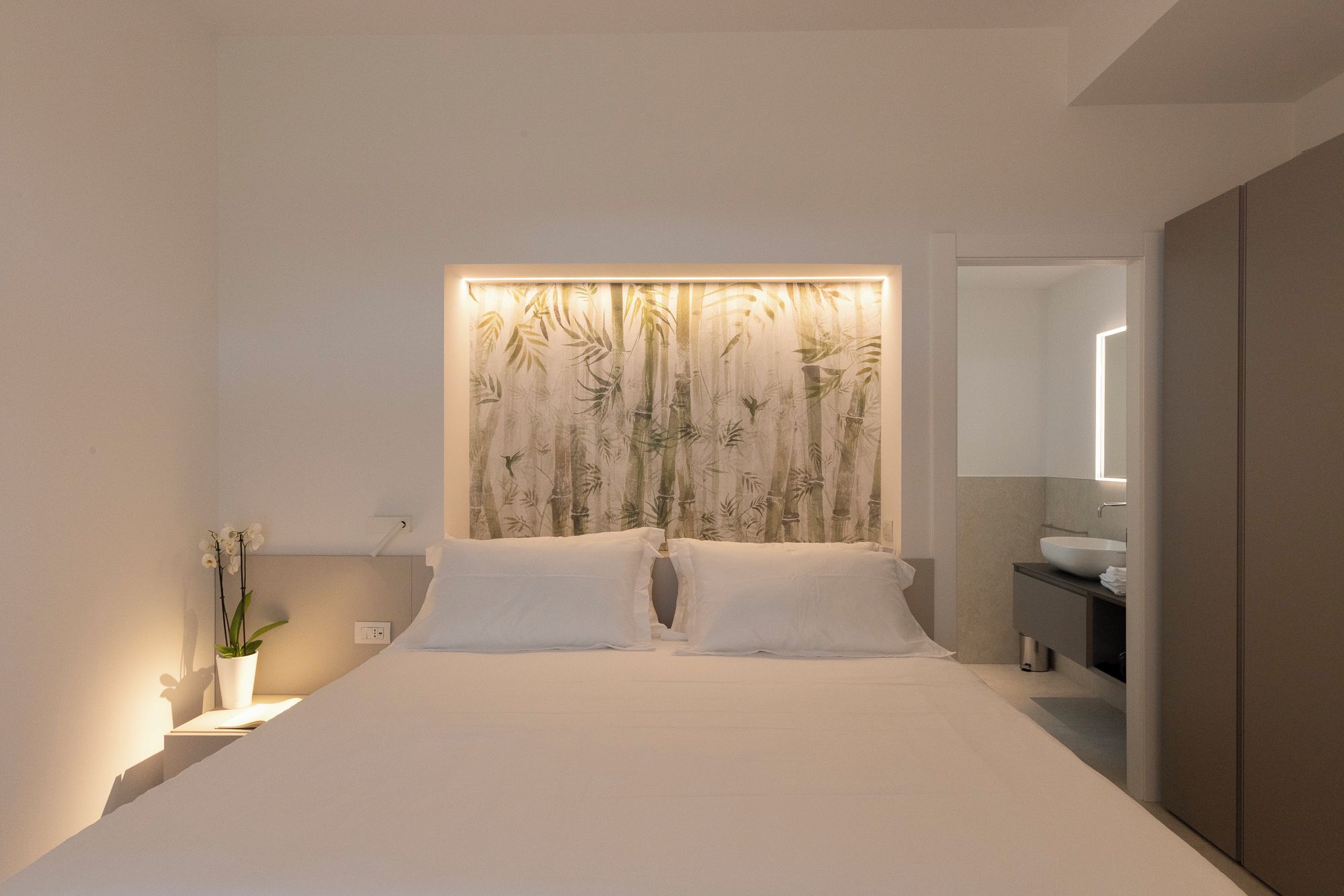 Un dormitorio moderno con una cama grande, cabecero de pared, mesitas de noche, una planta en maceta y un baño con lavamanos y espejo.