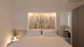 Un dormitorio moderno con una cama grande, cabecero de pared, mesitas de noche, una planta en maceta y un baño con lavamanos y espejo.