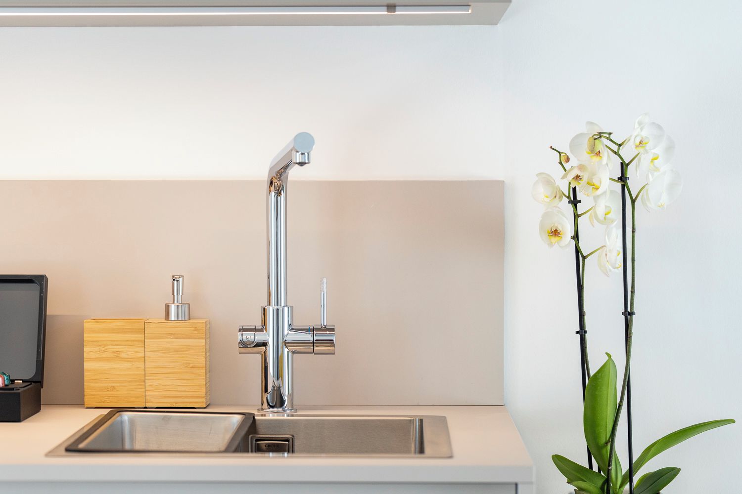 A modern kitchen with a stainless steel sink, wooden cabinet, and a potted orchid plant.