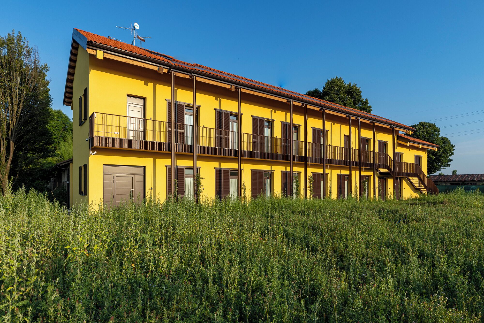 Un bâtiment jaune doté de nombreux balcons et surmonté d’un toit rouge, entouré de verdure.