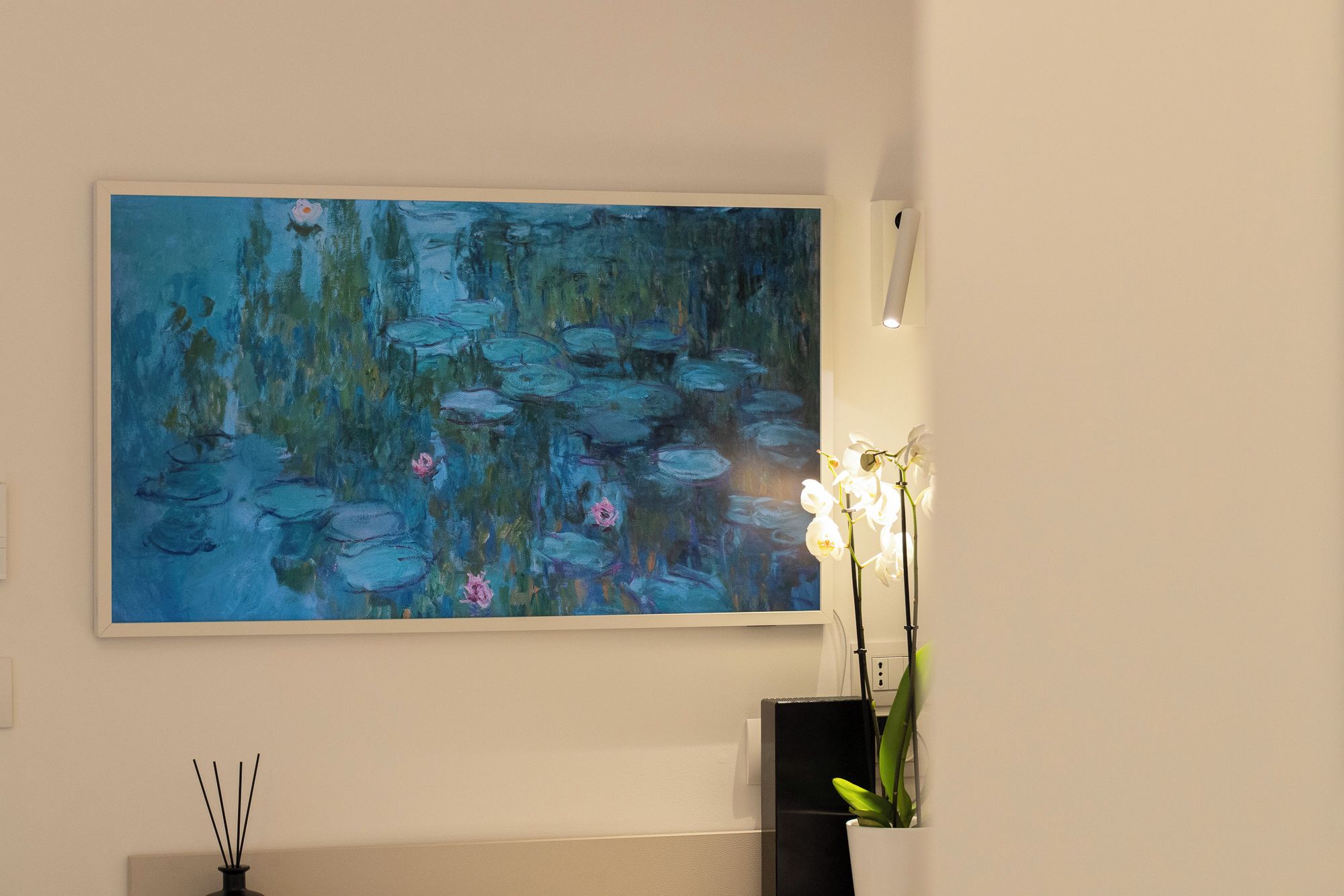 Un cuadro de nenúfares en una pared, un jarrón con flores blancas y un jarrón negro con hojas verdes.