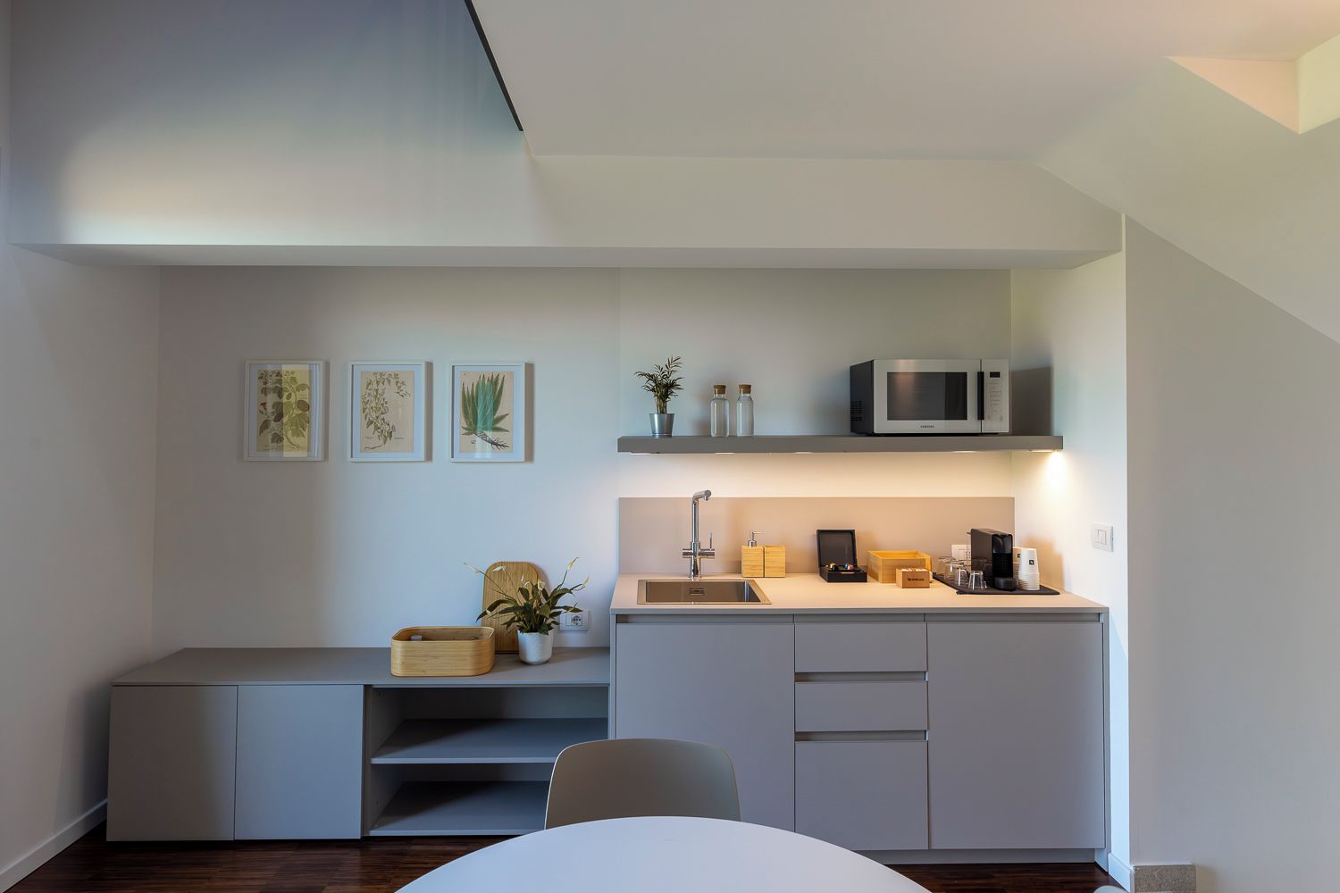 Cocina compacta con microondas, lavamanos y espacio de almacenamiento en la encimera. Hay grabados botánicos enmarcados en la pared y una pequeña planta en maceta sobre la encimera.