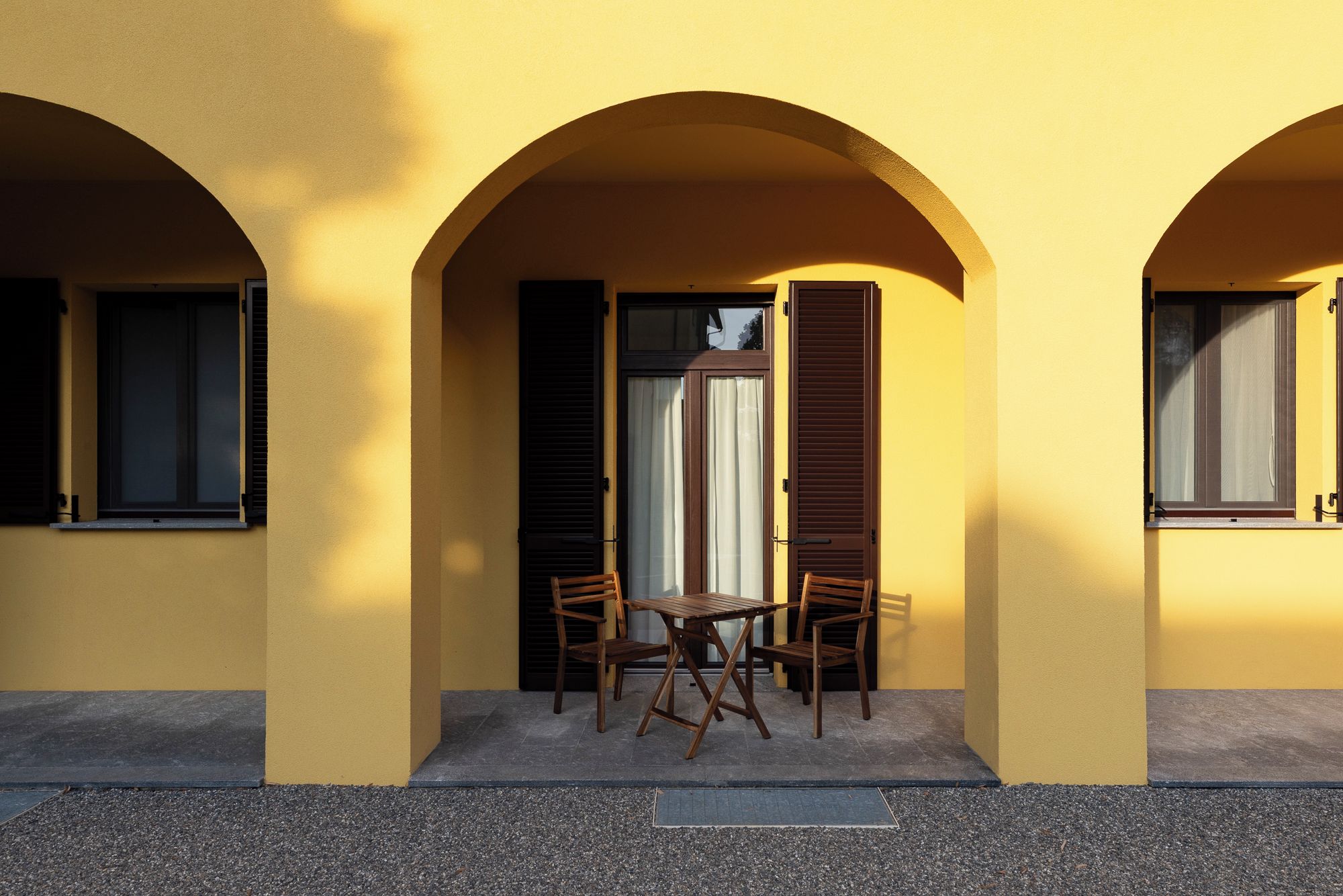 Un bâtiment doté de trois portes cintrées, chacune étant agrémentée d’une fenêtre et de volets. Une table et deux chaises se trouvent devant la porte centrale.