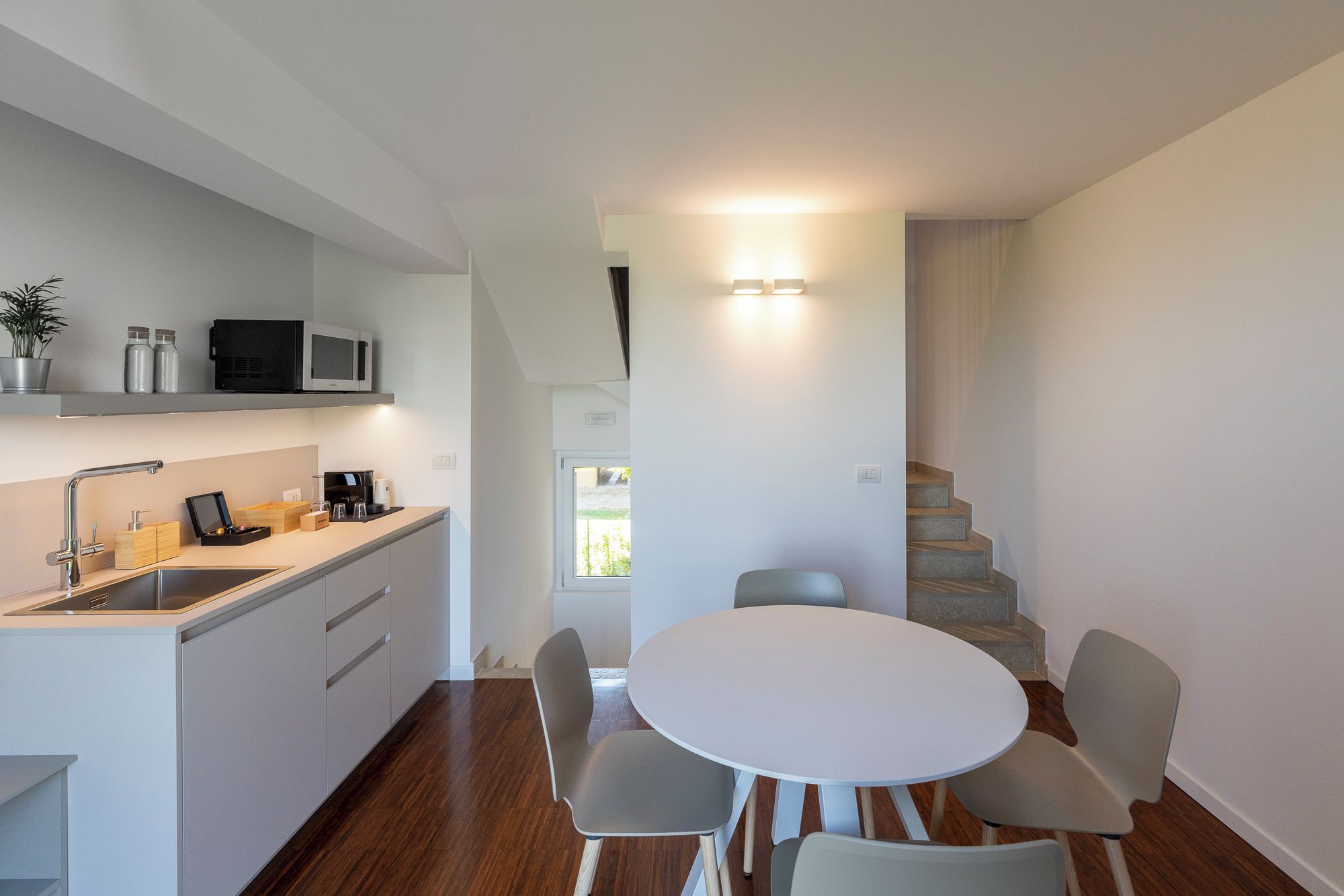 Una cocina compacta con encimera blanca, microondas y lavamanos. Hay un comedor con una mesa redonda y cuatro sillas. Se ve una escalera al fondo.