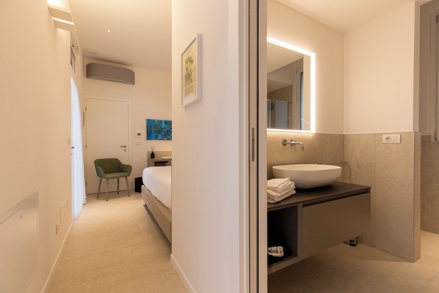Ett modernt hotellrum med en säng, en grön stol, ett badrum med en stor spegel och ett handfat.