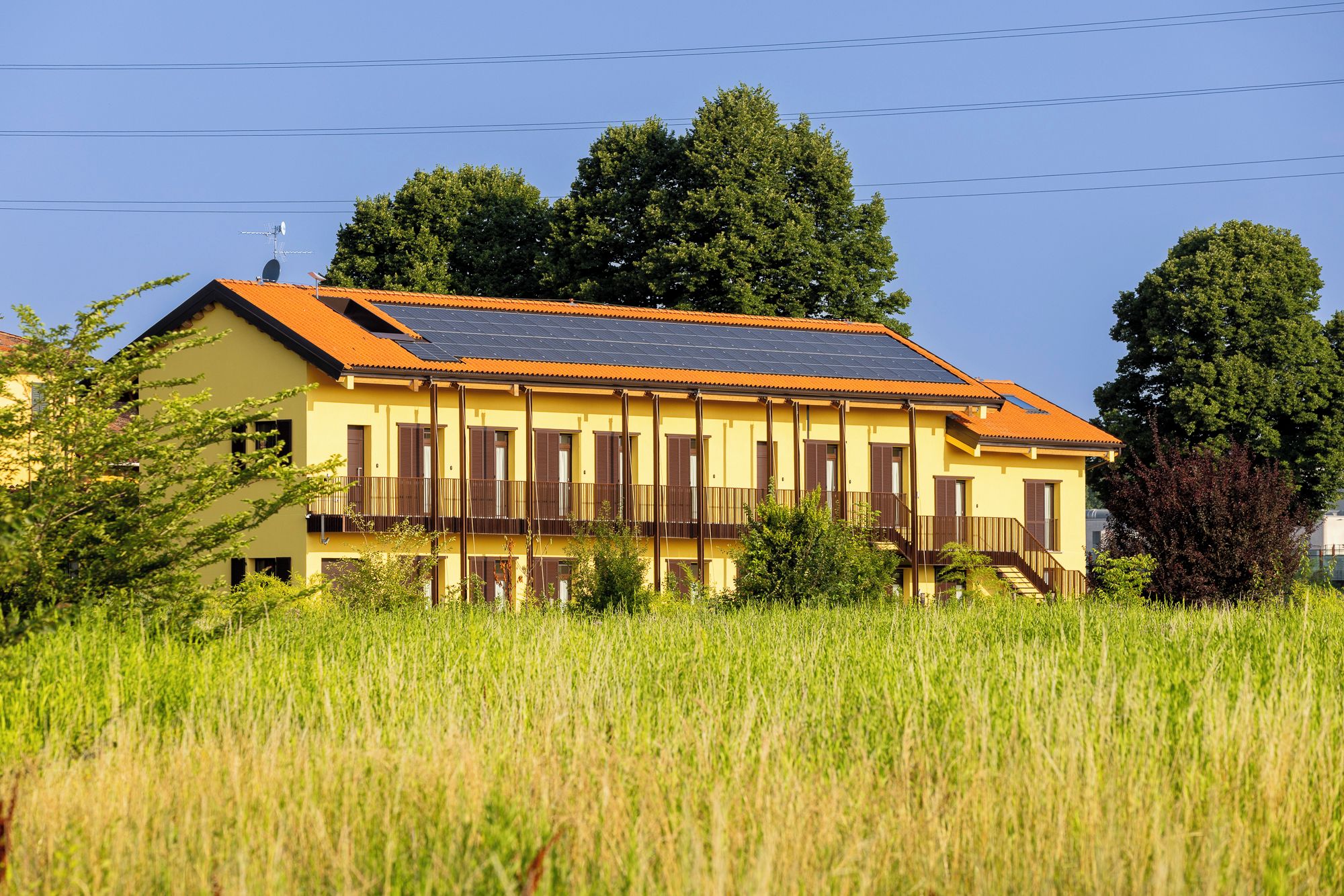 Un edificio amarillo con techo de paneles solares, rodeado de hierba alta y árboles.