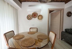 Un coin repas comprenant une table ronde en verre, quatre sets de table en fibres tressées, deux chaises et un climatiseur fixé au mur.