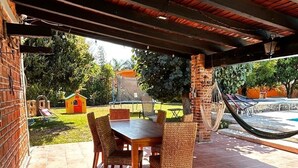 Un patio exterior con una mesa de madera y sillas de mimbre, una hamaca y una casita de juegos al fondo.