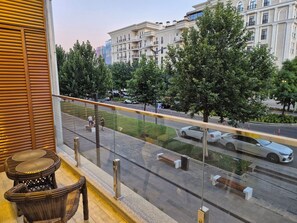 En balkon med fletningsmøbler, udsigt til en gade med biler, og en bygning med altaner i baggrunden.