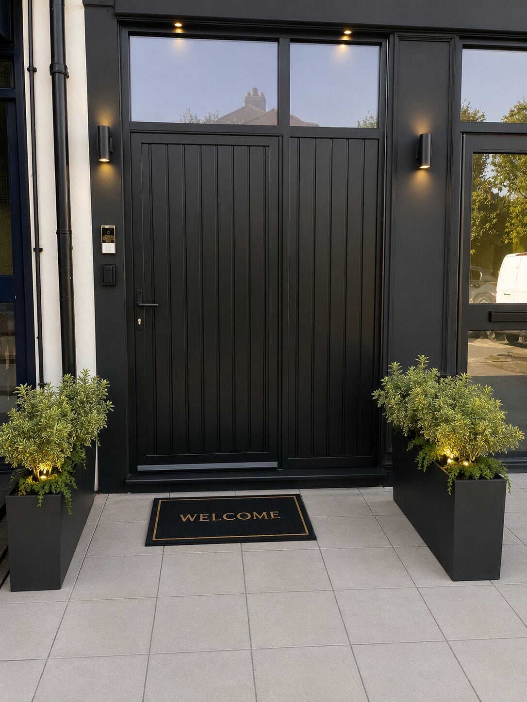A black double door with a 'WELCOME' mat, two potted plants, and a security system.