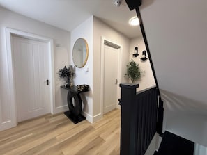 A hallway with a wooden floor, white doors, a black console table with decorative items, a round mirror, and a small potted plant. - Brand New Luxury 4 Bedroom 4 Bathroom Surbiton Flat | 
20min London Waterloo (Surbiton)