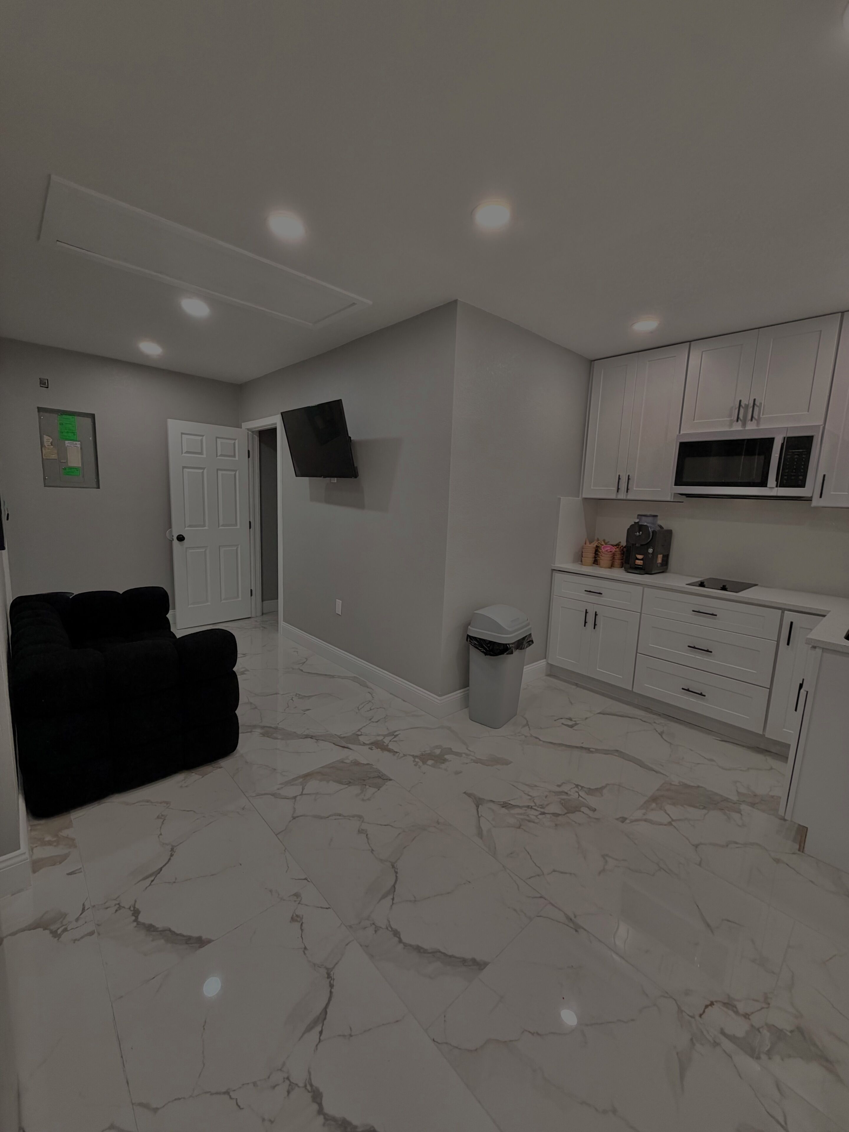 A modern living room with a black sofa, a television mounted on the wall, and a kitchen area with white cabinets and a microwave.