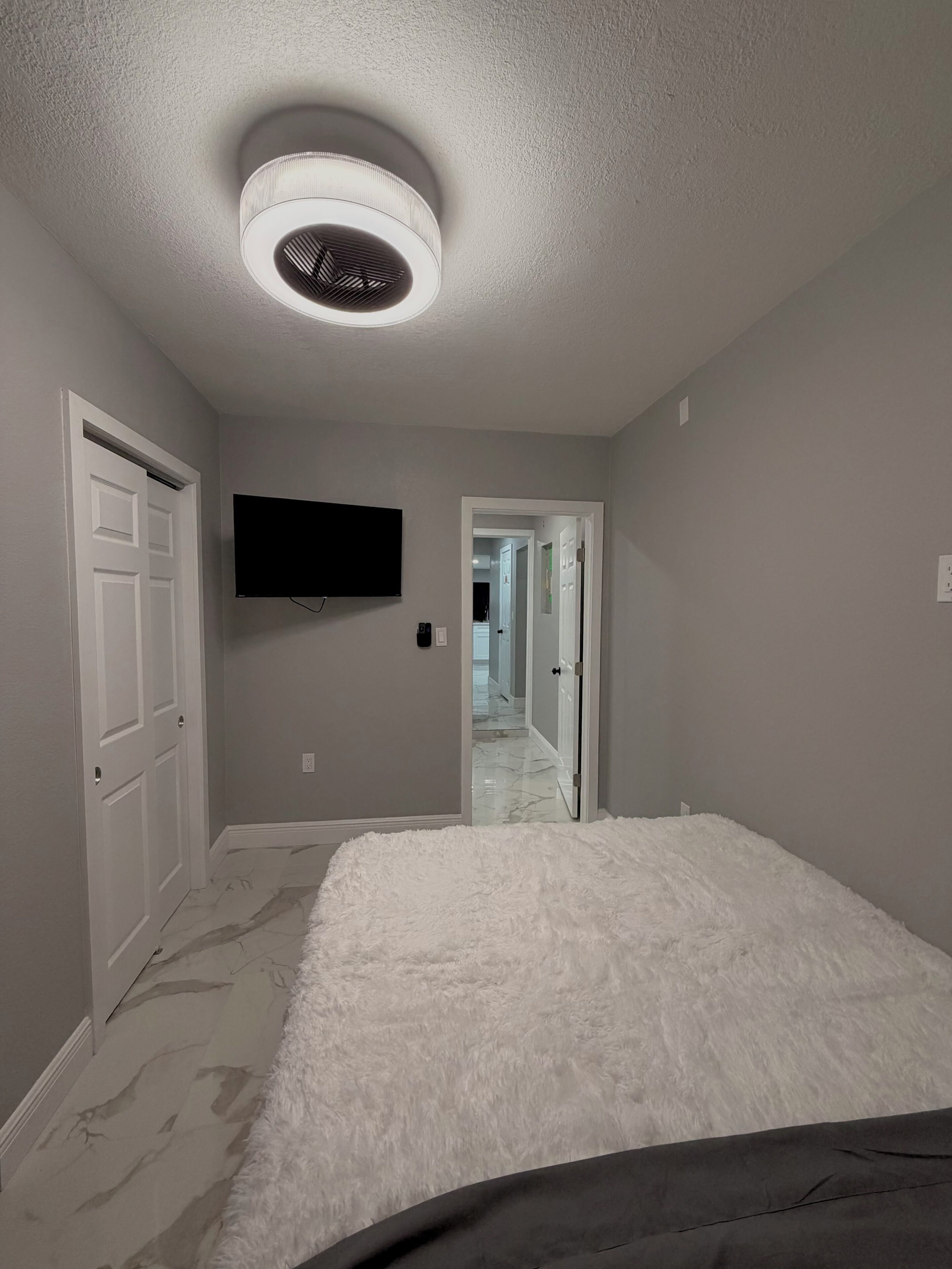 A bedroom with a bed, a ceiling fan, a TV, and a door leading to another room.
